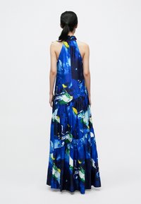 Maximekko, jossa on halterneck-mallinen kaula-aukko, eloisa sininen pohjaväri ja värikkäitä abstrakteja kuvioita sekä kerroksellinen hame. Sileä kangaspinta.