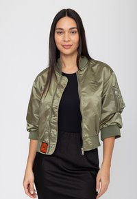 Giovane donna con capelli lunghi e scuri che indossa una giacca bomber verde oliva sopra un top e una gonna neri, in piedi contro uno sfondo chiaro e uniforme.