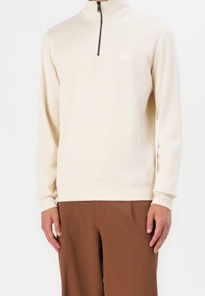 Mężczyzna ubrany w kremowy, długorękawowy pulower z czarnym zamkiem na ćwierć długości oraz brązowe spodnie, stojący na tle białej, gładkiej ściany.