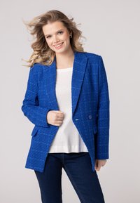 Modrý textúrovaný blazer s klopou, dvoma gombíkmi, prednými vreckami a voľným strihom, skombinovaný s bielym topom a tmavými džínsami.