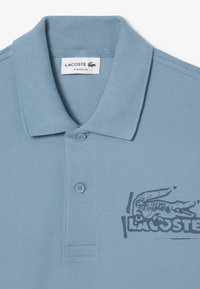 Jasnoniebieska koszulka polo wykonana z teksturowanej tkaniny, z dwoma guzikami przy kołnierzyku oraz nadrukowanym logo Lacoste w ciemnoniebieskim kolorze. Klasyczny krój.