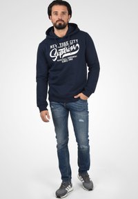 Marineblauwe hoodie met een voorste kangoeroezak en witte grafische tekst. Draagt bij gescheurde blauwe jeans en grijze sportschoenen.