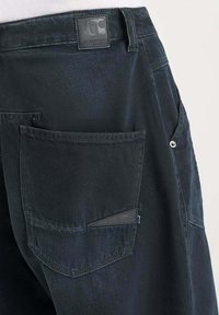 Zwarte denim broek met opvallende gestikte zakken, een leren label op de tailleband en een gestructureerde afwerking. Accenten omvatten een kleinere zak.