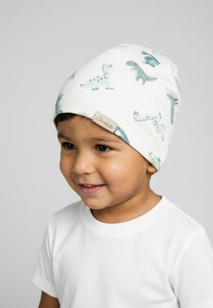 Young child wearing a white beanie with green dinosaur patterns and a white t-shirt, smiling and looking slightly to the side.