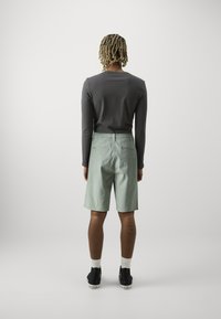 Pantalones cortos de color verde claro con corte recto y dos bolsillos traseros, combinados con una camiseta de manga larga ajustada de color gris oscuro. Zapatillas negras con suelas blancas.