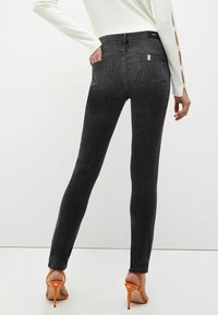 Nainen, joka nostaa tummaharmaita kapeita farkkuja ja oransseja korkokenkiä, seisoo yksi käsi takataskussa yksinkertaista taustaa vasten.