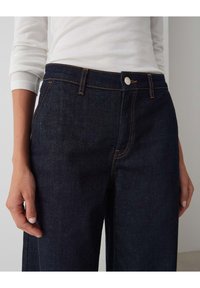 Dark blue denim jeans with a straight cut, featuring contrasting orange stitching and a silver metal button at the waist. Smooth texture.