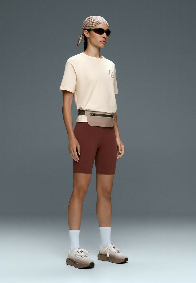 Personne portant un t-shirt beige, un short de cyclisme marron, des chaussettes blanches, des baskets beiges, des lunettes de soleil, un foulard et une sacoche beige sur fond gris.