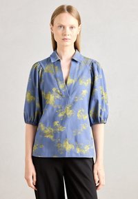 Modra bluzica z rumenim cvetličnim vzorcem, napihnjenimi rokavi in globokim V-izrezom. Lahka tkanina, ohlapen kroj, v kombinaciji s črnimi hlačami.