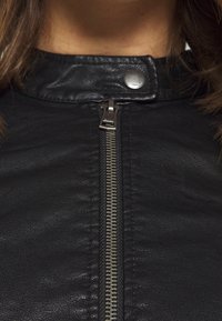 Giacca di pelle nera con colletto alto, zip in metallo argento e dettaglio di chiusura a scatto, caratterizzata da una texture liscia e cuciture ordinate.