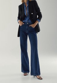 Blazer noir avec des boutons dorés, chemise en denim, jeans flare bleu taille haute et sandales à talons noires. Tissu lisse et coupe ajustée.