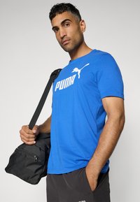 Zila kokvilnas T-krekls ar baltu "PUMA" logotipu un grafiku uz krūtīm. Modelis tur melnu somu, valkājot tumšas šortus ar kabatām.
