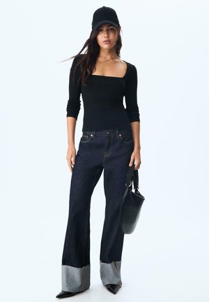 Haut noir à manches longues avec décolleté carré, associé à un jean large bleu foncé avec des accents de poignet gris. Un sac noir et un chapeau complètent le look.