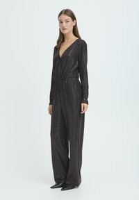 Czarny kombinezon z długim rękawem, głębokim dekoltem w serek, elastyczną talią i teksturowanym wzorem. Noszony z czarnymi butami o szpiczastym nosku.