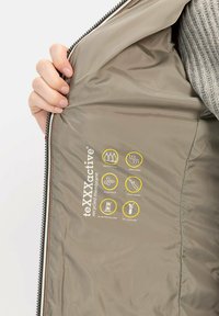 Gerecycled membraanjack in beige, met een gladde textuur. Aan de binnenkant zijn gele iconen te zien voor waterdicht, ademend, winddicht en andere functies.