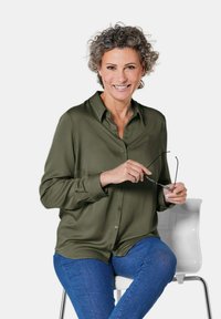 Olivgrüne Bluse mit Knopfleiste und Kragen, lange Ärmel; getragen mit blauen Jeans. Person hält eine Brille und sitzt auf einem weißen Stuhl.