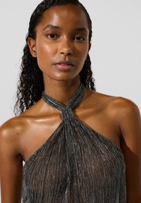 Melns haltertops no caurspīdīga, krokaina auduma ar metālisku rotājumu. Kakla izgriezums priekšpusē ir ar mezgla dizainu.