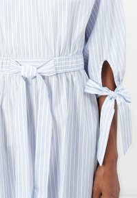 Vestido listrado em azul claro e branco, com cinto na cintura e laços nas mangas, revelando a pele nas fendas, exibido em uma pessoa com tom de pele de média-escuridão.