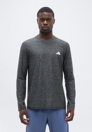 Mann trägt ein dunkelgraues langärmeliges Sportshirt mit weißem Logo auf der Brust und blaue Shorts, steht vor einem einfarbigen Hintergrund.