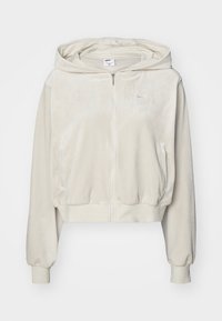 Krátká mikina s kapucí z měkké světle béžové látky, se zipem vpředu, kapucí se šňůrkou a mírně nafouklými rukávy. Na hrudi je malá logo.