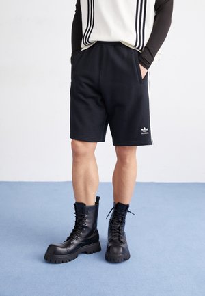 Mężczyzna w czarnych szortach Adidas i czarnych masywnych butach sznurowanych, stojący na jasnoniebieskiej podłodze z rękami w kieszeniach.