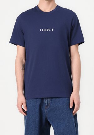 Person trägt ein dunkelblaues T-Shirt mit weißem Text "JORDAN" in auseinander gesetzten Buchstaben und locker sitzende blaue Jeans vor weißem Hintergrund.