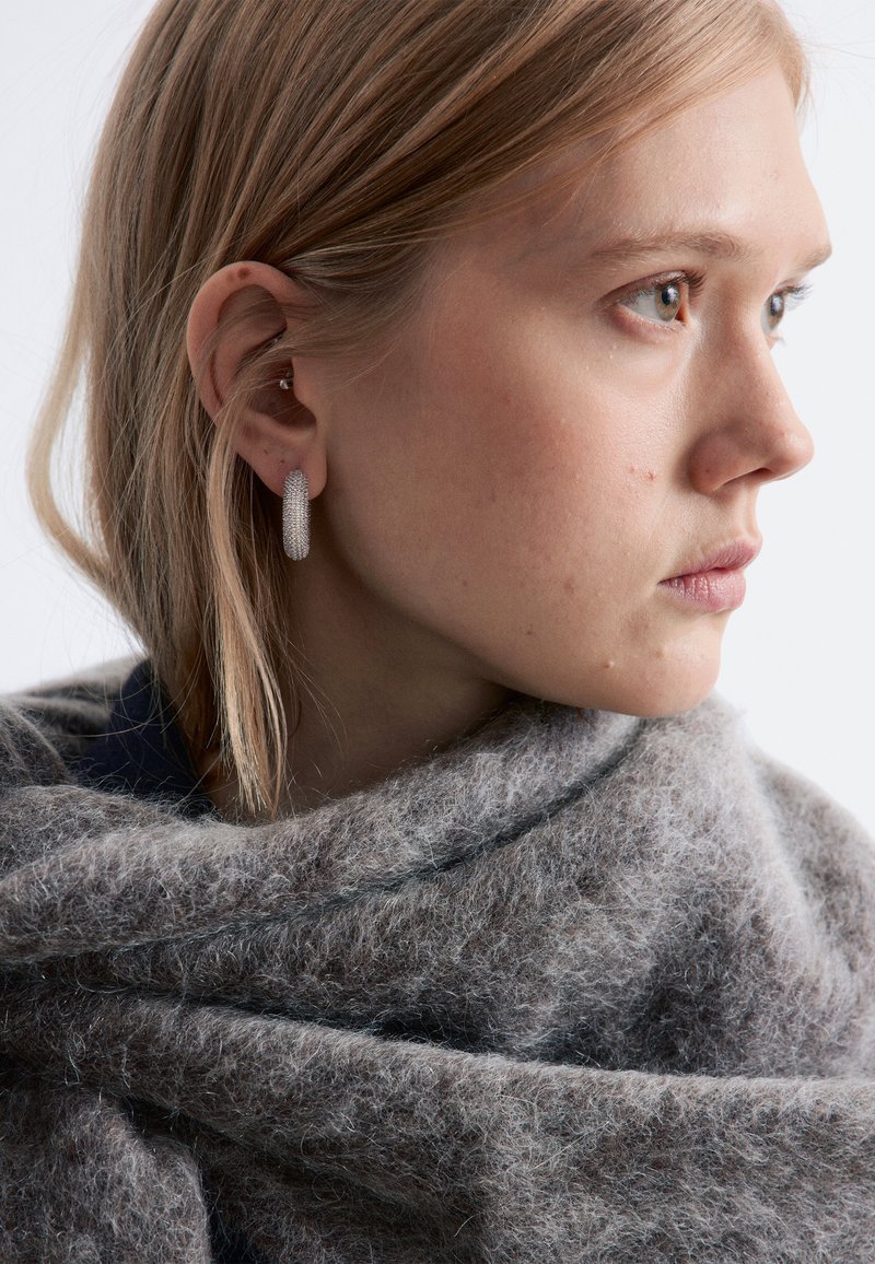 Boucles d'oreilles créoles en argent texturé portées par une personne drapée dans une écharpe en laine grise douce, avec des fibres et des plis visibles.