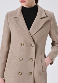 Cappotto beige doppiopetto realizzato in tessuto caldo, con colletto a rever, due tasche frontali e bottoni dorati per la chiusura.