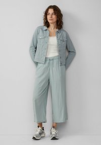 Hellblaue Denimjacke über einem weißen Tanktop, kombiniert mit lockeren hellblauen Hosen und silbernen Sportschuhen.