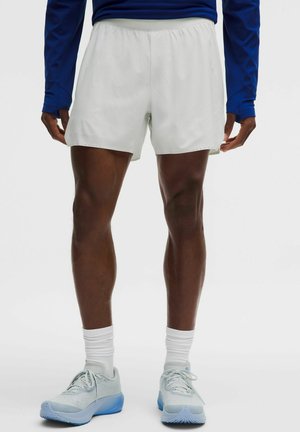 Man die witte sportshorts, witte sportsokken, lichtblauwe en witte hardloopschoenen en een donkerblauw shirt met lange mouwen draagt, staand.