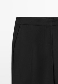 Schwarze, maßgeschneiderte Hose mit glattem Bund, scharfen Bügelfalten, Seitentaschen und strukturierter Textur. Das Design besticht durch einen sauberen, modernen Schnitt.