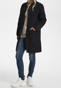 Abrigo largo negro acolchado con cierre de cremallera, bolsillos laterales y puños acanalados, llevado sobre una blusa ligera de satén y jeans ajustados, combinado con zapatillas blancas.