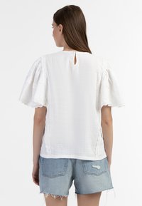 Hvid bomuldsbluse med puffærmer, bølget blonderegning langs siderne og en lille nøglehulrygdetalje. Kombineret med denimshorts.