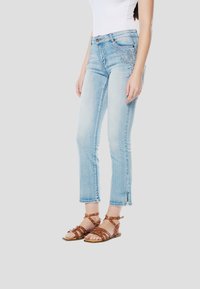 Lichtblauwe spijkerbroek met rechte pasvorm, voorzien van geborduurde bloemmotieven en rafelige zoom, gecombineerd met bruine sandaaltjes met studs.