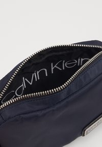 Granatowa saszetka Calvin Klein z metalowym zamkiem błyskawicznym, częściowo otwarta, ukazująca czarne wnętrze wykończone białym tekstem Calvin Klein.