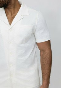 Chemise blanche à manches courtes avec un motif texturé, design à col, une seule poche sur la gauche et boutons blancs. Tissu léger.