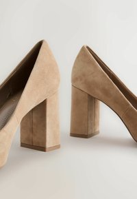 Paire d’escarpins beige en daim à talons blocs avec bouts pointus sur un fond neutre.