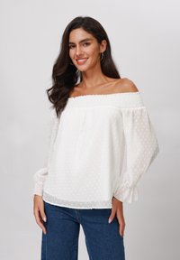 White off-shoulder blouse with textured polka dots, smocked neckline, sheer long sleeves, and elastic cuffs. Paired with blue jeans.