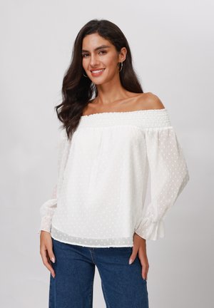 Blouse blanche à épaules dénudées avec des pois texturés, encolure smockée, manches longues transparentes et poignets élastiques. Assortie avec un jean bleu.
