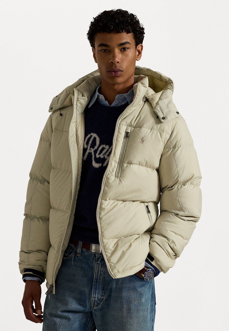 Light beige puffer jacket with a hood, zippered front, quilted design, side pockets, and embroidered logo on the chest. Worn over a sweater.