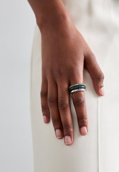 Anillo de plata y verde oscuro colocado en una mano, con una banda lisa y acentos texturizados, y dos anillos apilados, descansando sobre una superficie de color claro.