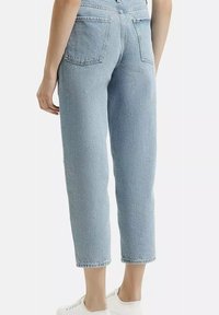 Osoba ubrana w jasnoniebieskie dżinsy z wysokim stanem i proste nogawki oraz białe trampki, pokazana od tyłu na jednolitym tle.