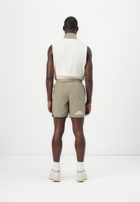 Haut sans manches blanc avec col beige, assorti à un short vert olive arborant un logo discret. Le mannequin porte des baskets claires et des chaussettes blanches.
