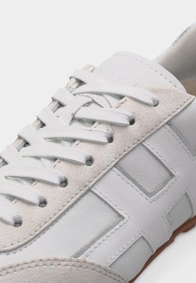 Άσπρο δερμάτινο και σουέντ αθλητικό παπούτσι με λευκά κορδόνια και ραμμένες γεωμετρικές επενδύσεις, με καφέ καουτσούκ περίγραμμα στην σόλα.