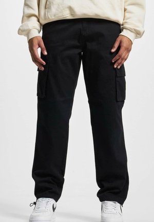 Persona con pantalones cargo negros con bolsillos laterales y zapatillas blancas, de pie frente a un fondo liso.