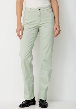 Frau trägt hellgrüne, gerade geschnittene Hosen und schwarze Loafers, steht vor einem schlichten weißen Hintergrund.