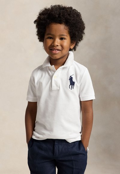 Jeune garçon souriant aux cheveux bouclés portant un polo blanc avec un logo de cheval et un pantalon foncé, debout les mains dans les poches.