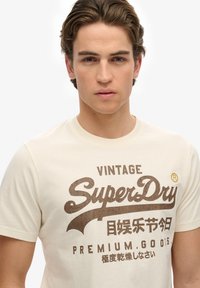 Beige bomulds t-shirt med korte ærmer, der har "Vintage SuperDry" logo i brunt, akkompagneret af japanske tegn og et buet design.