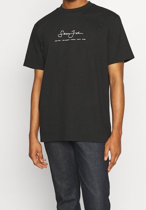 Mand iført sort Sean John t-shirt med korte ærmer, hvidt logo tekst, kombineret med mørkeblå jeans, stående mod en ensfarvet baggrund.