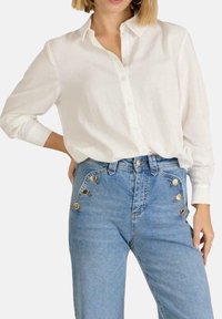 Camisa blanca de manga larga y botones hecha de una tela ligera, combinada con vaqueros azules de cintura alta con acentos de botones metálicos.